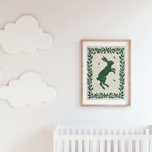 May include: A green rabbit silhouette print with a leafy border, framed in natural wood. The print is hanging on a white wall with white cloud-shaped decorations.