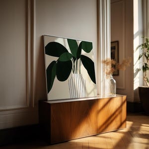 May include: A framed art print featuring a stylized green plant in a striped vase. The artwork is displayed on a wooden cabinet, with a clear glass vase and a potted plant in the background. The room has a warm, natural light.