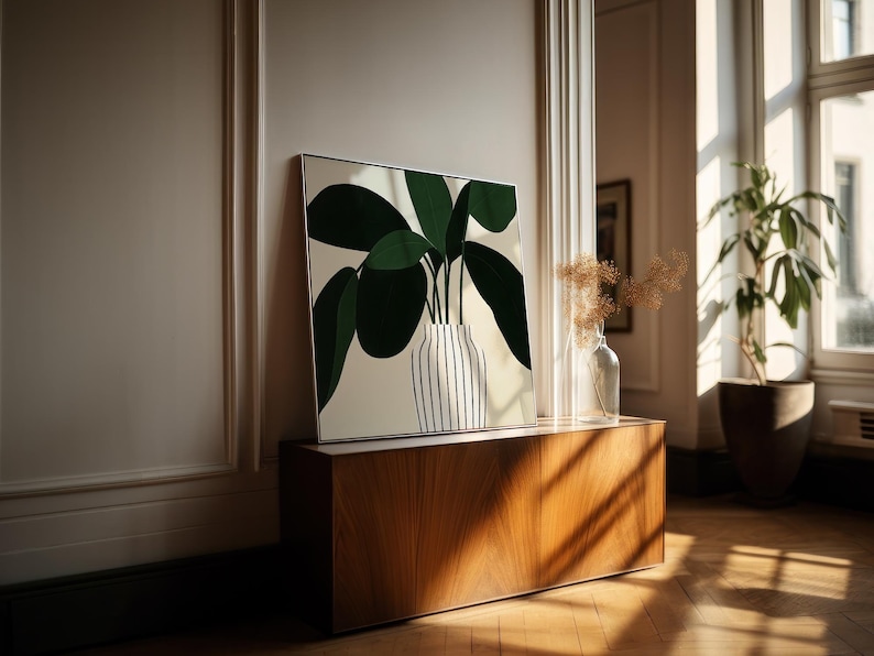 May include: A framed art print featuring a stylized green plant in a striped vase. The artwork is displayed on a wooden cabinet, with a clear glass vase and a potted plant in the background. The room has a warm, natural light.