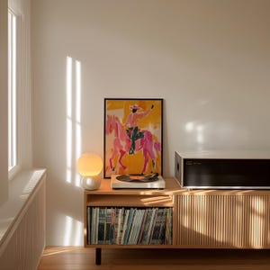 Pu&ograve; includere: Un giradischi in stile moderno di met&agrave; secolo con un giradischi bianco e un sistema di altoparlanti nero si trova su un mobile in legno con uno scaffale di dischi in vinile. Una lampada a globo bianca si trova sul mobile accanto al giradischi. Una stampa incorniciata di un cavallo rosa e un cavaliere &egrave; appesa al muro dietro il mobile.