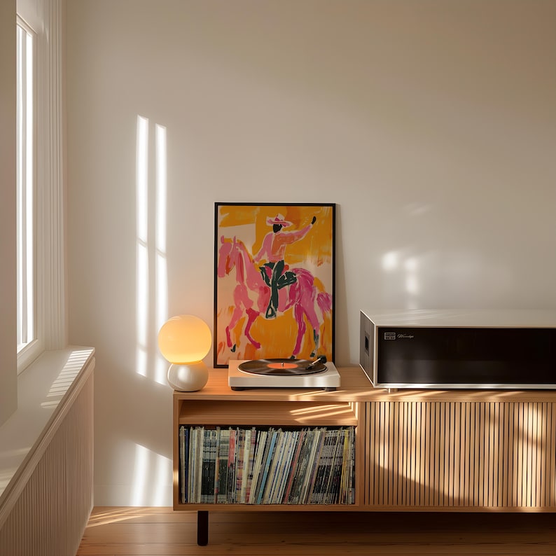 Pu&ograve; includere: Un giradischi in stile moderno di met&agrave; secolo con un giradischi bianco e un sistema di altoparlanti nero si trova su un mobile in legno con uno scaffale di dischi in vinile. Una lampada a globo bianca si trova sul mobile accanto al giradischi. Una stampa incorniciata di un cavallo rosa e un cavaliere &egrave; appesa al muro dietro il mobile.