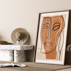 Op de afbeelding: Een ingelijst abstract lijntjeskunstprint van een gezicht in bruin en beige tinten. De print hangt aan een beige muur met een houten vloer en een witte terrazzo bank op de voorgrond.