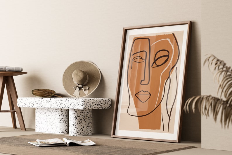Op de afbeelding: Een ingelijst abstract lijntjeskunstprint van een gezicht in bruin en beige tinten. De print hangt aan een beige muur met een houten vloer en een witte terrazzo bank op de voorgrond.