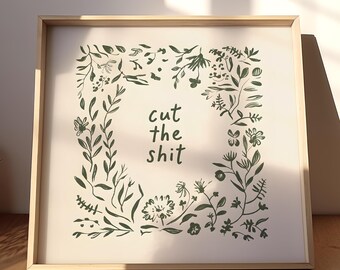 May include: Square framed art print with a light beige background. The print features a dark green floral border surrounding the words "cut the shit" in a handwritten style font. The frame is a light wood color.