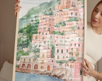 Puede incluir: Una pintura de acuarela de un pueblo costero con edificios rosas y amarillos, árboles verdes y un océano azul. La pintura está enmarcada en un marco de madera claro.