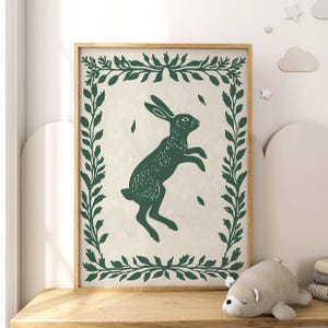 May include: A green and white print of a rabbit jumping in a leafy frame. The rabbit is silhouetted in dark green against a cream background.