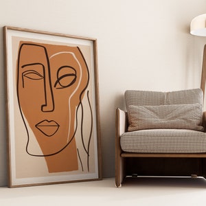 Op de afbeelding: Een ingelijst abstract lijntjeskunstprint van een gezicht in bruin en zwart op een beige achtergrond. De print hangt aan een witte muur naast een bruine fauteuil met een geruit kussen.