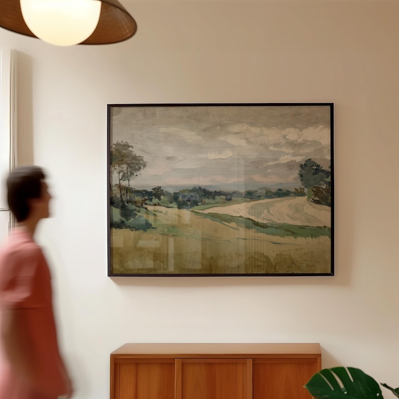 K&ouml;nnte beinhalten: Ein gerahmtes Landschaftsgem&auml;lde zeigt eine l&auml;ndliche Szene mit Feldern, B&auml;umen und einem bew&ouml;lkten Himmel. Das Kunstwerk verwendet ged&auml;mpfte T&ouml;ne von Gr&uuml;n, Beige und Grau und erzeugt eine ruhige und naturalistische &Auml;sthetik. Das Gem&auml;lde wird an einer wei&szlig;en Wand ausgestellt.