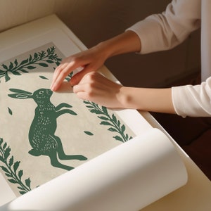May include: A green and white print of a rabbit hopping with green foliage surrounding it. The print is on a white background and is being rolled up.