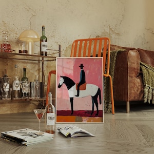 K&ouml;nnte beinhalten: Eine gerahmte Illustration einer Person, die auf einem wei&szlig;en und schwarzen Pferd auf rosafarbenem Hintergrund reitet. Die Illustration ist in einem goldenen Rahmen und liegt auf einem Holzboden.