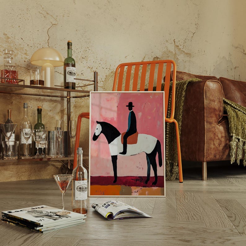 K&ouml;nnte beinhalten: Eine gerahmte Illustration einer Person, die auf einem wei&szlig;en und schwarzen Pferd auf rosafarbenem Hintergrund reitet. Die Illustration ist in einem goldenen Rahmen und liegt auf einem Holzboden.