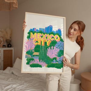 Könnte beinhalten: Ein gerahmter Kunstdruck mit der Skyline von Mexiko-Stadt in Blau über einer lebendigen Landschaft aus grünen Kakteen, rosa Wüstenflora und dem gelben Schriftzug MEXICO CITY. Die Illustration verwendet einen kräftigen, farbenfrohen Grafikstil.