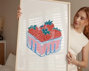 Può includere: Una stampa incorniciata con una vivace illustrazione di fragole in un cestino rosa e blu. Le fragole sono rosse con le foglie verdi e il cestino ha un contorno blu. L'opera d'arte è su sfondo bianco.