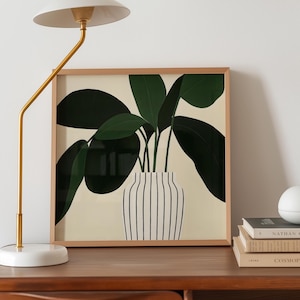May include: A framed art print featuring a stylized illustration of a plant with large, dark green leaves. The plant is in a white vase with vertical black stripes, set against a cream background. The frame is made of light-colored wood.