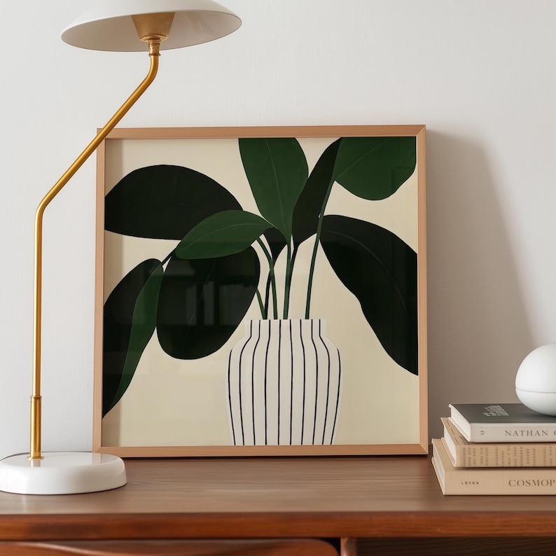 May include: A framed art print featuring a stylized illustration of a plant with large, dark green leaves. The plant is in a white vase with vertical black stripes, set against a cream background. The frame is made of light-colored wood.