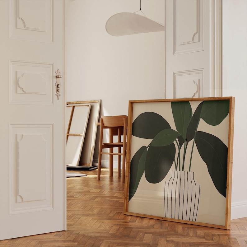 May include: A framed botanical print featuring a stylized illustration of a plant with large, dark green leaves in a striped vase. The artwork is set against a neutral background and framed in natural wood. The print leans against a wall in a room with hardwood floors.