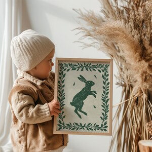 May include: A child holds a framed print of a green rabbit silhouette against a white background. The print has a green leafy border and the rabbit is jumping in the air.