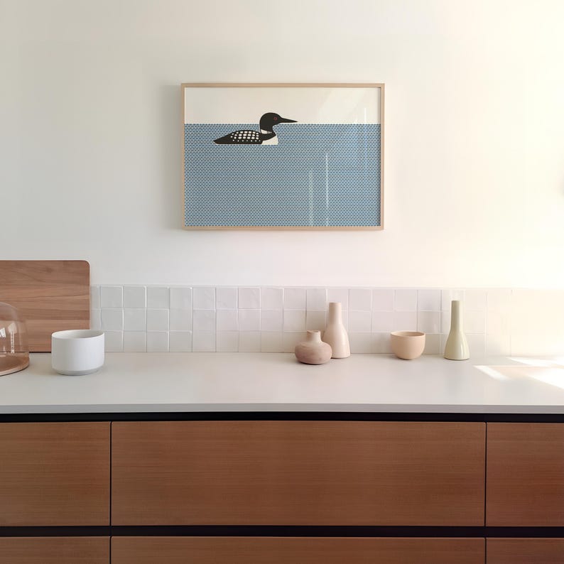 May include: A framed art print featuring a minimalist illustration of a loon swimming in blue water. The artwork is displayed above a countertop with decorative vases and bowls. The cabinet doors are made of wood.