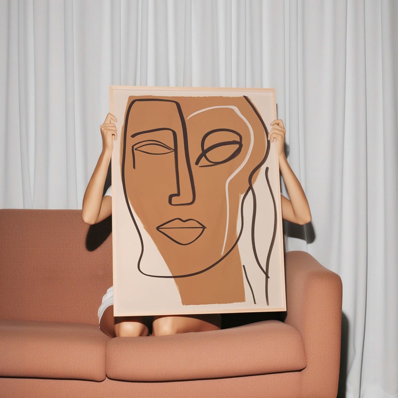 Op de afbeelding: Een persoon houdt een ingelijst abstract kunstprint vast met een bruin en beige gezicht met zwarte lijnen. De print is op een witte achtergrond.