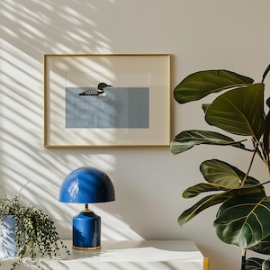 May include: A framed art print featuring a loon swimming in blue water. The artwork is in a gold frame and hangs above a blue table lamp with a gold base. A potted plant is on the left, and a large-leafed plant is on the right.