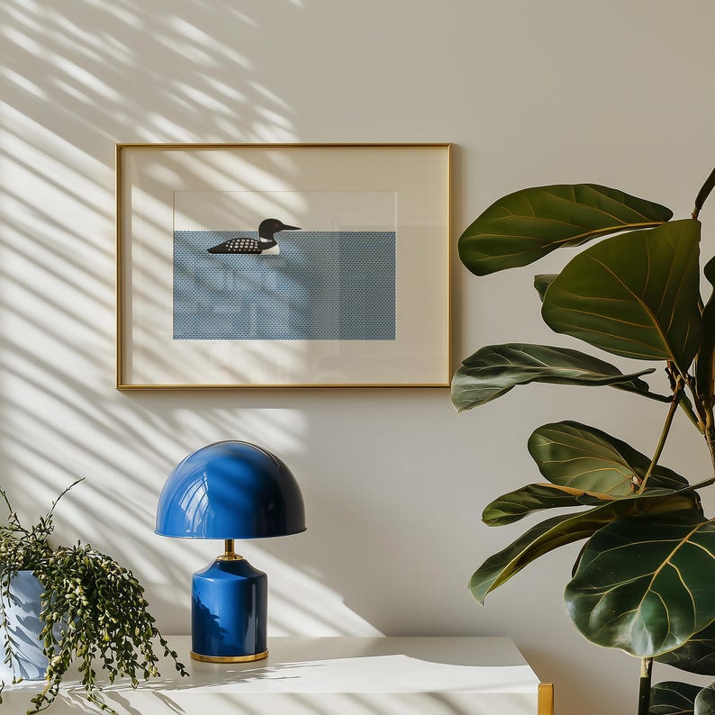 May include: A framed art print featuring a loon swimming in blue water. The artwork is in a gold frame and hangs above a blue table lamp with a gold base. A potted plant is on the left, and a large-leafed plant is on the right.