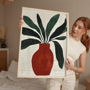 May include: A framed illustration of a red vase with green leaves. The vase is a simple, rounded shape and the leaves are stylized and abstract.