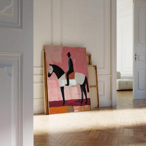 K&ouml;nnte beinhalten: Ein gerahmtes Gem&auml;lde, das ein schwarz-wei&szlig;es Pferd mit einer Person in einem schwarzen Hut und einem blauen Hemd zeigt, die darauf reitet. Der Hintergrund ist ein abstraktes Design in Rosa und Orange.