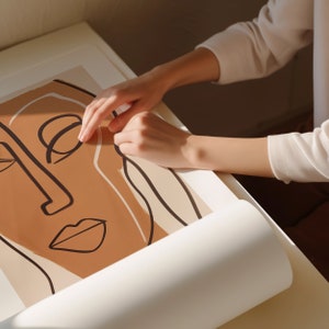 Op de afbeelding: Een persoon rolt een afdruk van een bruin en wit abstract gezicht met zwarte lijnen op. De afdruk is op wit papier.