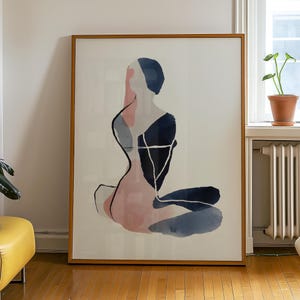 May include: Abstract art print featuring a seated figure in shades of pink, navy, and gray. The artwork is framed in a natural wood tone and hangs on a white wall. The figure's form is suggested with watercolor-like brushstrokes.