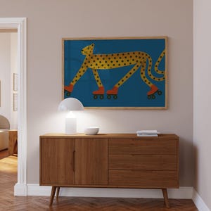 May include: A framed art print shows a yellow cheetah with black spots wearing orange roller skates on a blue background. The artwork hangs above a mid-century modern wooden cabinet with a white lamp and bowl.