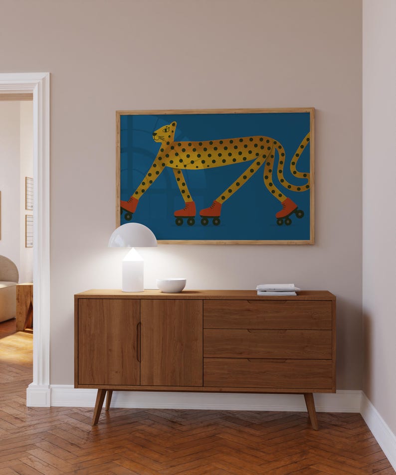 May include: A framed art print shows a yellow cheetah with black spots wearing orange roller skates on a blue background. The artwork hangs above a mid-century modern wooden cabinet with a white lamp and bowl.