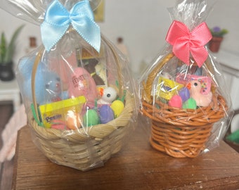 May include: Two miniature Easter baskets filled with colorful treats. The basket on the left has a blue bow and contains a pink bunny, chocolate eggs, and a yellow box of Peeps. The basket on the right has a pink bow and contains a pink chick, chocolate eggs, and a purple egg.