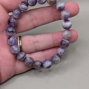 May include: A beaded bracelet featuring round, polished amethyst stones. The bracelet's beads display a mix of purple and white hues, with natural patterns. The bracelet is held in a hand, showcasing the gemstone's color and texture.