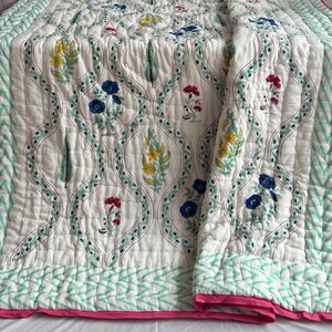 Può includere: Una trapunta bianca trapuntata con un motivo floreale blu, giallo e rosso. La trapunta ha un bordo a zig-zag verde e bianco e un bordo rosa. Il design presenta un motivo ripetuto di fiori e foglie stilizzate.