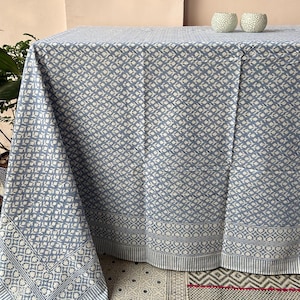 May include: A blue and white patterned tablecloth. The tablecloth has a repeating floral design and a border with a geometric pattern. The fabric is draped over a table, with a portion of the tablecloth hanging down. Two small white decorative objects are on the table.