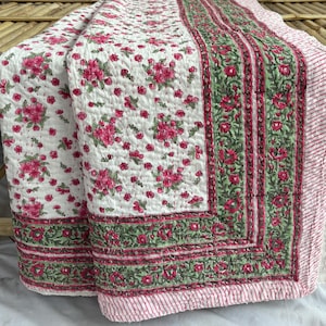 Puede incluir: Un edredón floral blanco con un estampado de flores rosas y detalles verdes. El edredón tiene un borde a rayas en rosa y blanco. El edredón está doblado sobre un banco de madera.