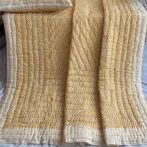 Pode incluir: Uma colcha com padrão chevron amarelo mostarda e branco com duas almofadas a condizer. As almofadas têm um fundo branco com um padrão chevron amarelo mostarda e uma borda decorativa. A colcha e as almofadas são feitas de tecido acolchoado macio.