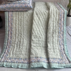 May include: A light green quilted bedspread and two matching pillowcases. The bedding features a floral pattern with blue and pink accents. The edges of the bedspread and pillowcases are trimmed with a green and white striped border. The bedding is arranged on a bed.