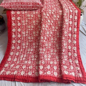 May include: A red and white floral patterned quilt with matching pillow shams. The bedding set features a repeating floral design in white against a red background. The quilt and pillow shams are neatly arranged, showcasing the intricate pattern and color scheme.
