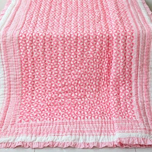 May include: A rectangular quilted blanket featuring a repeating pink and white geometric pinwheel pattern. The centre is framed by a white border and a striped pink and white edge, finished with a ruffled trim along all four sides of the textile.