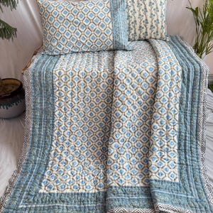 以下が含まれることがあります： 青と白の幾何学模様の綿のキルト。キルトは白と黒の幾何学模様の青い縁取りがあります。キルトは半分に折り畳まれ、白い表面の上に置かれています。