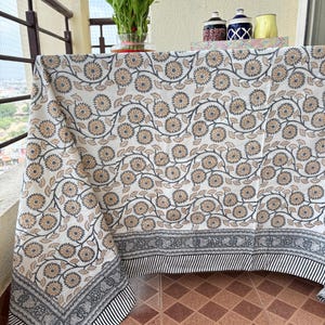 May include: A white tablecloth with a repeating floral pattern in shades of tan and gray. The border features a black and white striped design with a decorative gray pattern. The tablecloth is draped over a table on a balcony.