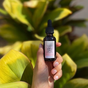 May include: A small, dark brown glass bottle with a dropper top. The bottle is labeled "Nona Alchemy Botanica" and "PMS Support". The label also features a butterfly and crescent moon design.