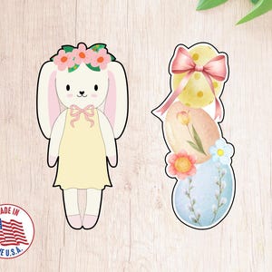 Può includere: Due formine per biscotti a tema pasquale. Uno è un coniglietto con una corona di fiori e un vestito. L'altro è una pila di uova decorate con un fiocco e fiori. Adesivo Made in the U.S.A.