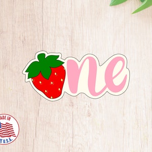May include: Pink and white "one" with a red and green strawberry graphic. The graphic is on the left side of the word "one".