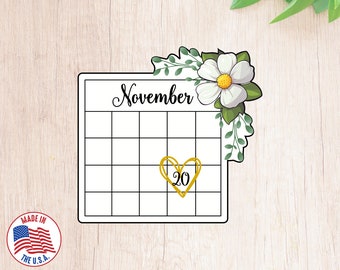 May include: A white calendar with a floral design in the top right corner. The calendar is for November and has a gold heart around the number 20.