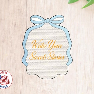 Puede incluir: Un letrero decorativo azul y blanco con un lazo en la parte superior. El letrero tiene un fondo texturizado y el texto "Write Your Sweet Stories" en naranja.