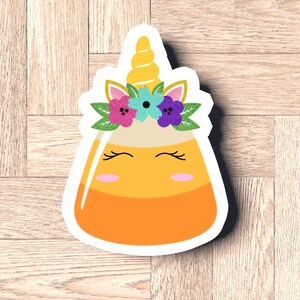 Könnte beinhalten: Ein gelber und orangefarbener Maisbonbon mit einem Einhornhorn und einer Blumenkrone. Das Einhorn hat ein lächelndes Gesicht mit geschlossenen Augen und rosa Wangen.
