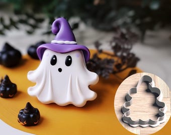 Può includere: Un tagliapasta a forma di fantasma bianco con un cappello da strega viola e un'espressione sorpresa. Il tagliapasta è su una superficie gialla con decorazioni nere e marroni.
