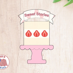 May include: A pink cake stand with a white cake on top. The cake is decorated with three red strawberry designs and a banner that says "Sweet Stories".